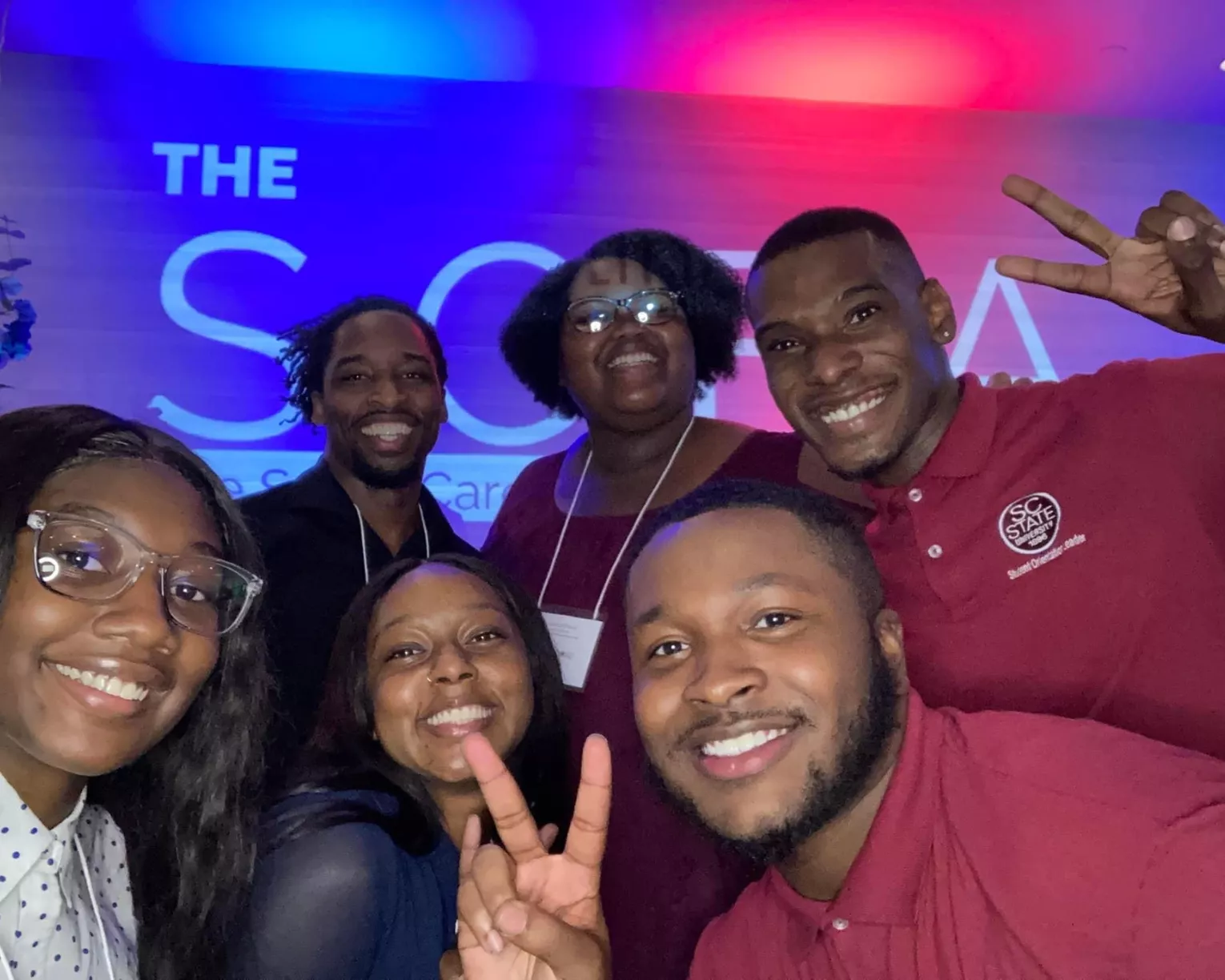 Six aspiring educator members from SC State University pose for a photo. Three student members are in the front, all are smiling, and the student in the right corner is flashing a peace sign. There are three students in the back row, also smiling, with the person to the furthest right flashing a peace sign. In the background, there are blue and pink neon lights and the image of The SCEA logo in white is just barely visible in the spaces between people.