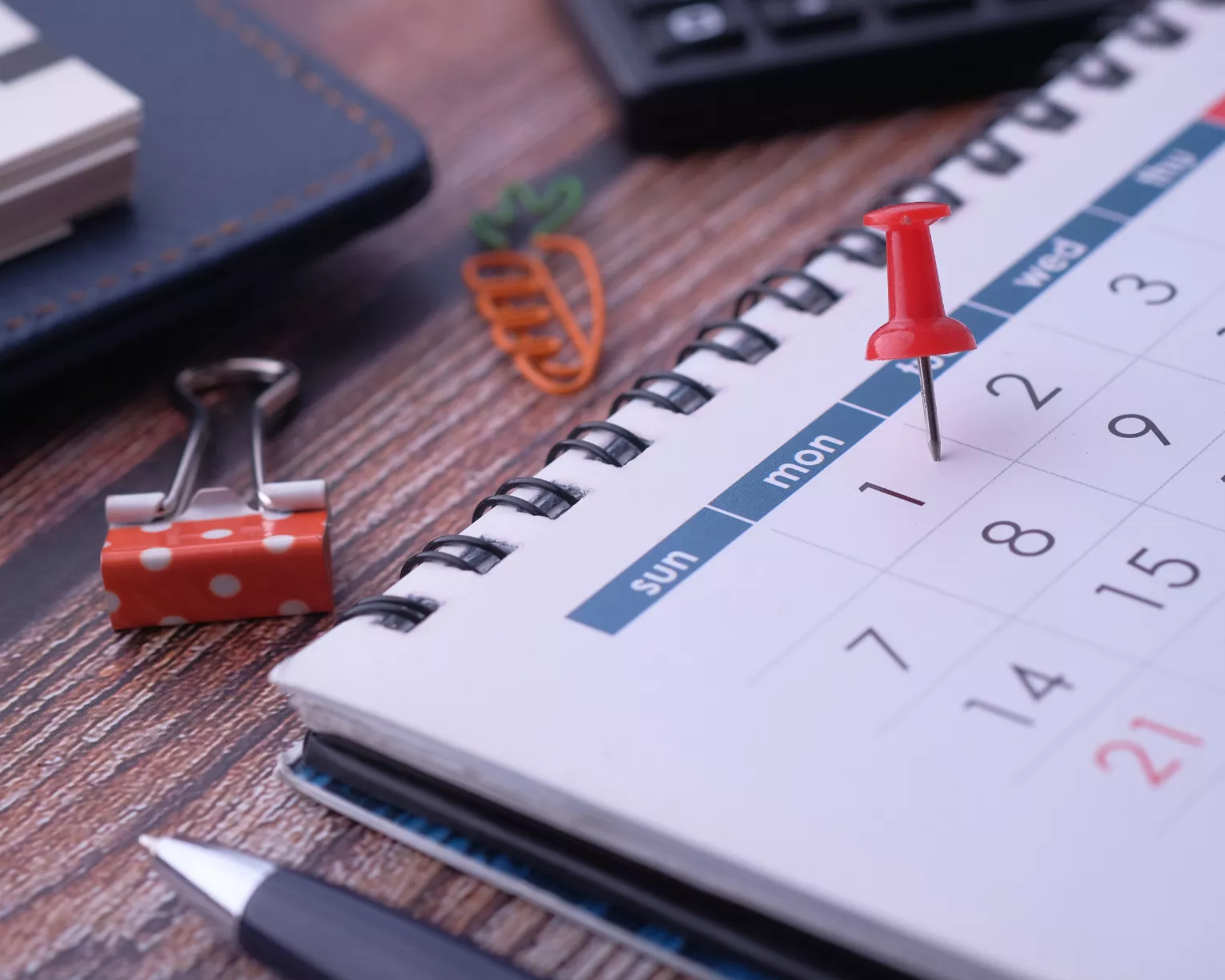 A calendar in a planning book sits on a wooden desk with a red tac pushed into the 2nd.