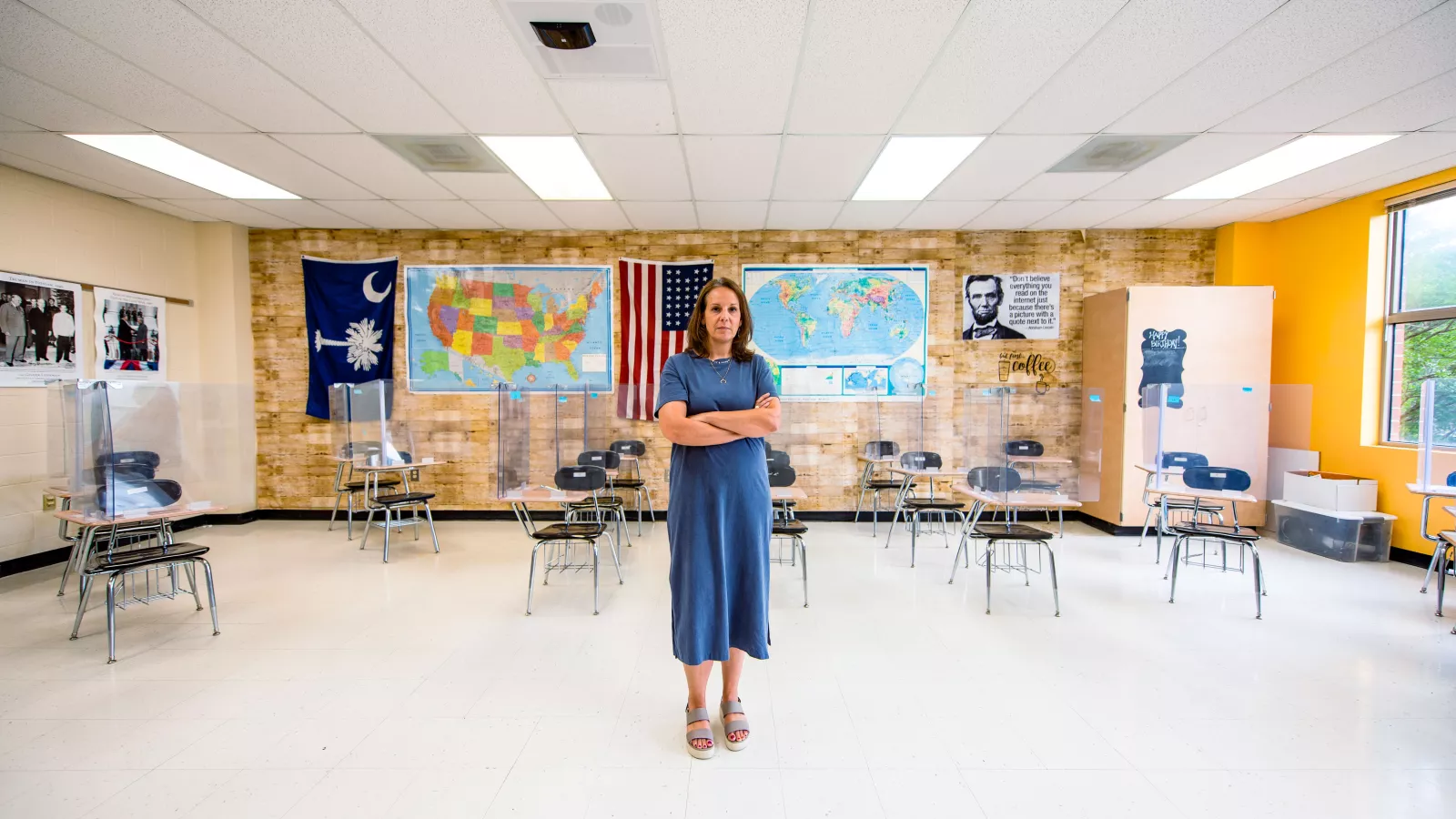 Teacher stands with arms crossed in a Social Studies classroom with plexiglass on desks.