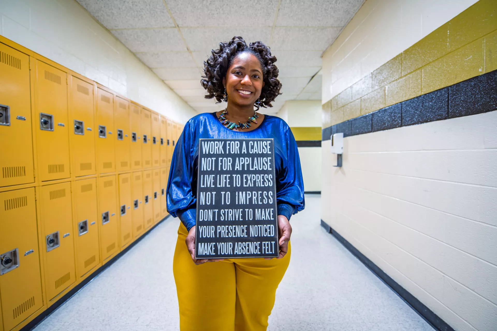 A teacher poses in a school hallway with a sign about leadership.