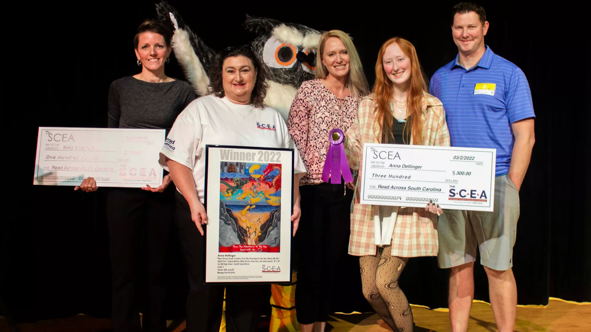 The art contest winner poses with The SCEA President Sherry East, The SCEA mascot Ollie the Owl, her art teacher, and her family. Pictured left to right: Art teacher Kelly Lucarelli, The SCEA President Sherry East, Anna Dellinger's mother, student artist Anna Dellinger, Anna Dellinger's father. Lucarelli and Dillinger are both holding large checks, and Sherry East is holding a print of Dellinger's award winning art.