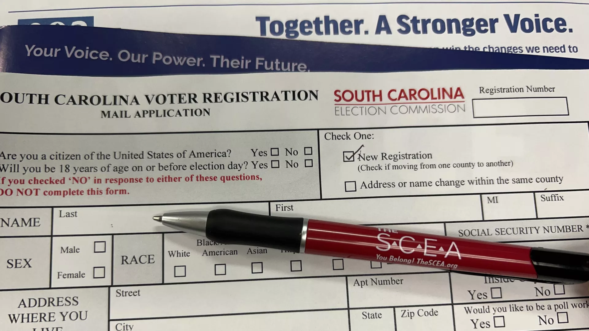 A red pen with "The SCEA" on the pen lies on top of a blank South Carolina voter registration form.