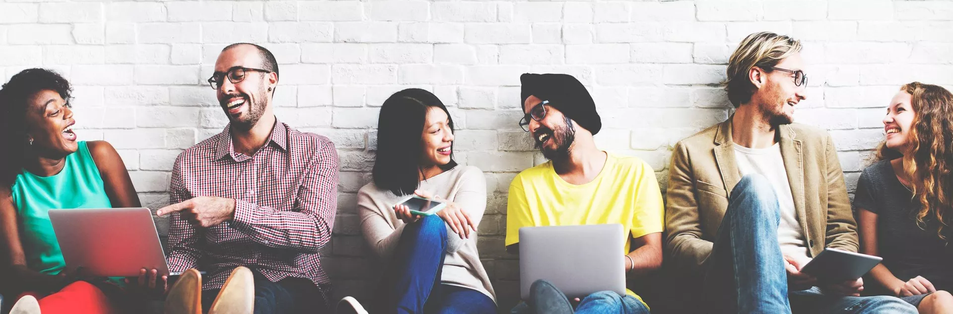 Six people sit against a white brick wall. They are laughing and talking. Two of the people have laptops they are sharing with the others.