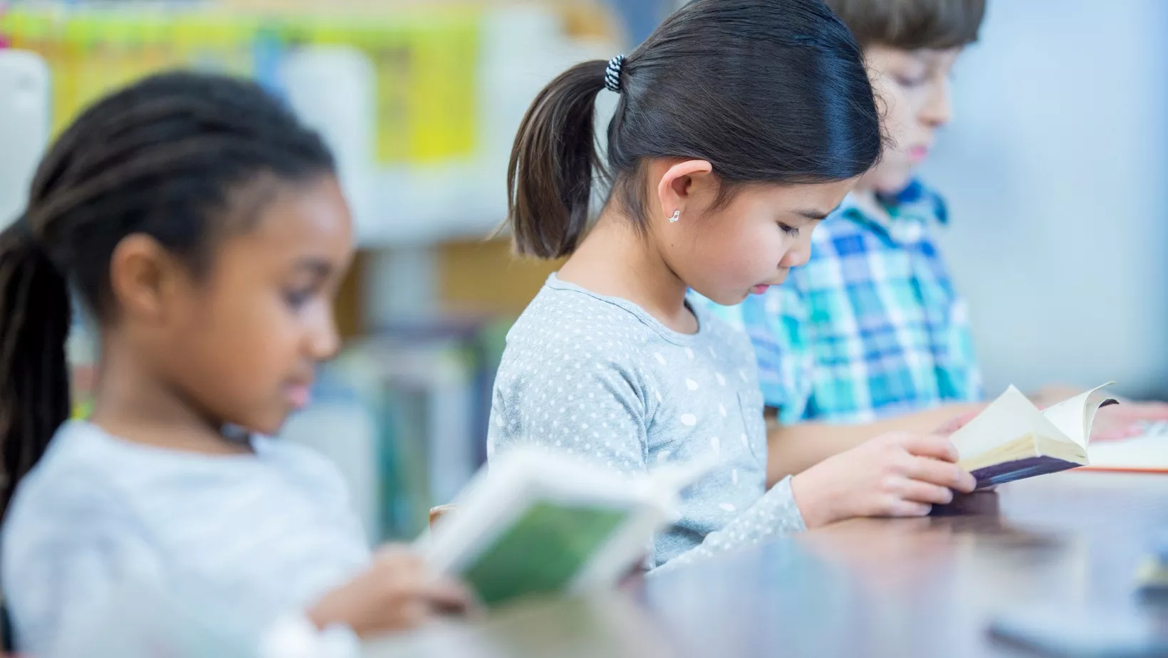 Three children sit at a table. Each child is reading a book.