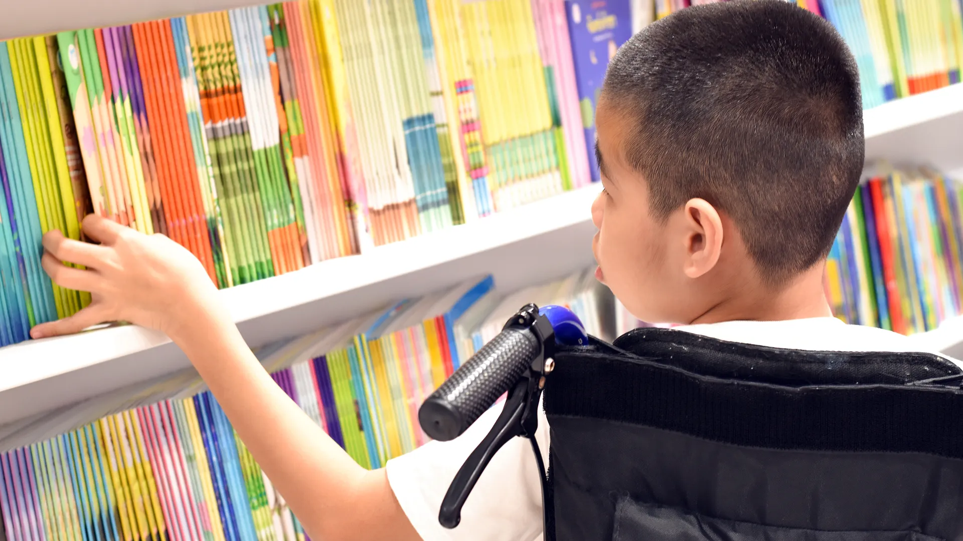 young asian boy in a wheelchair chooses books in library
