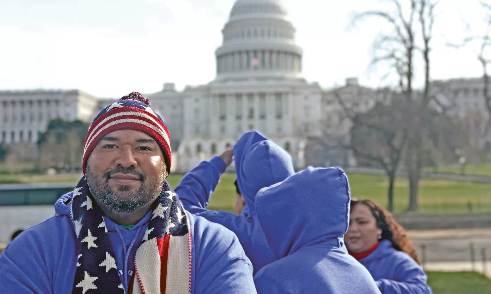 Jorge Botella stands outside in Washington DC with an American flag scarf and hat and the US capitol building in the background