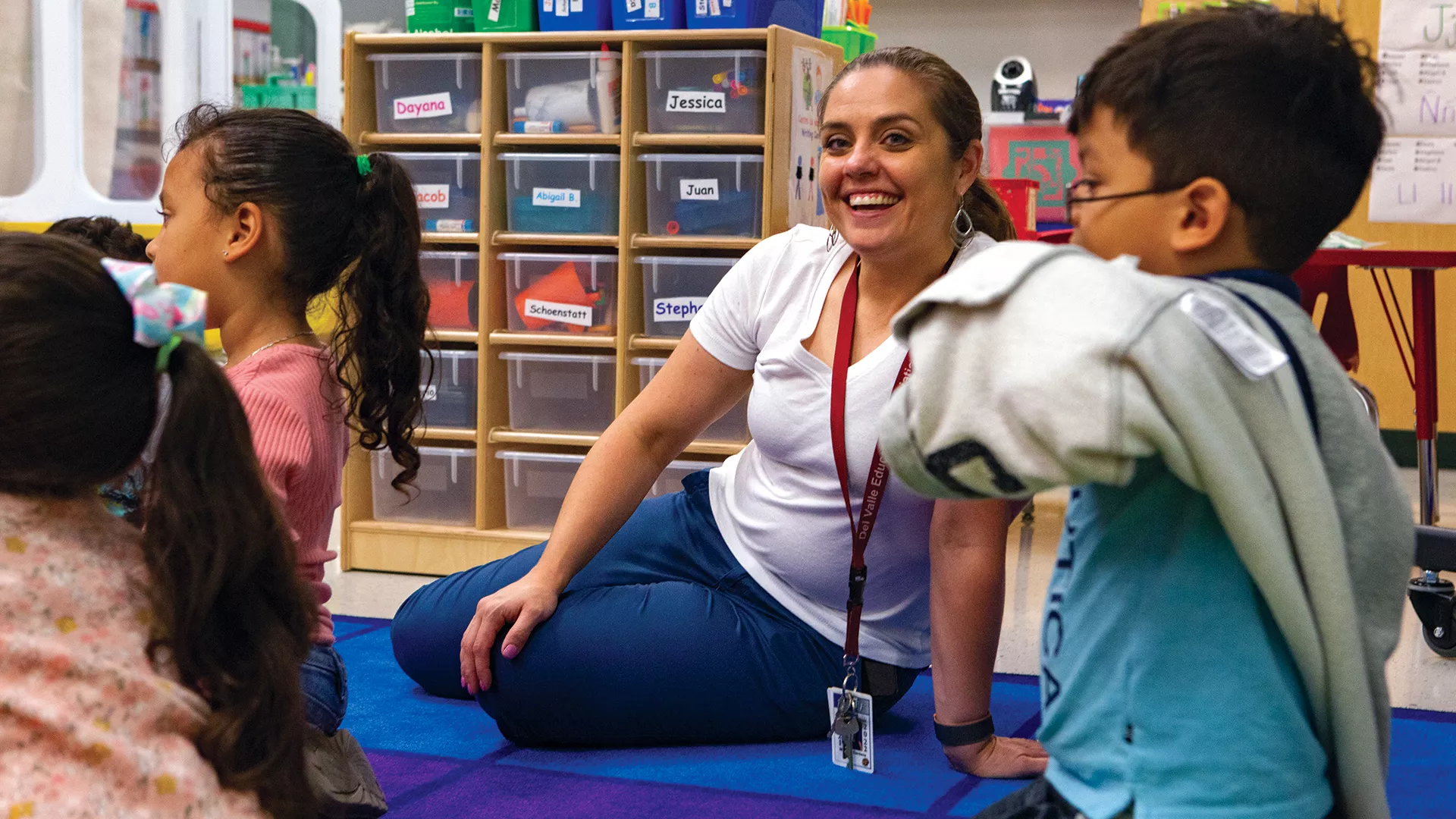 Teacher Michelle Cardenas sits on a rug in her classroom with students