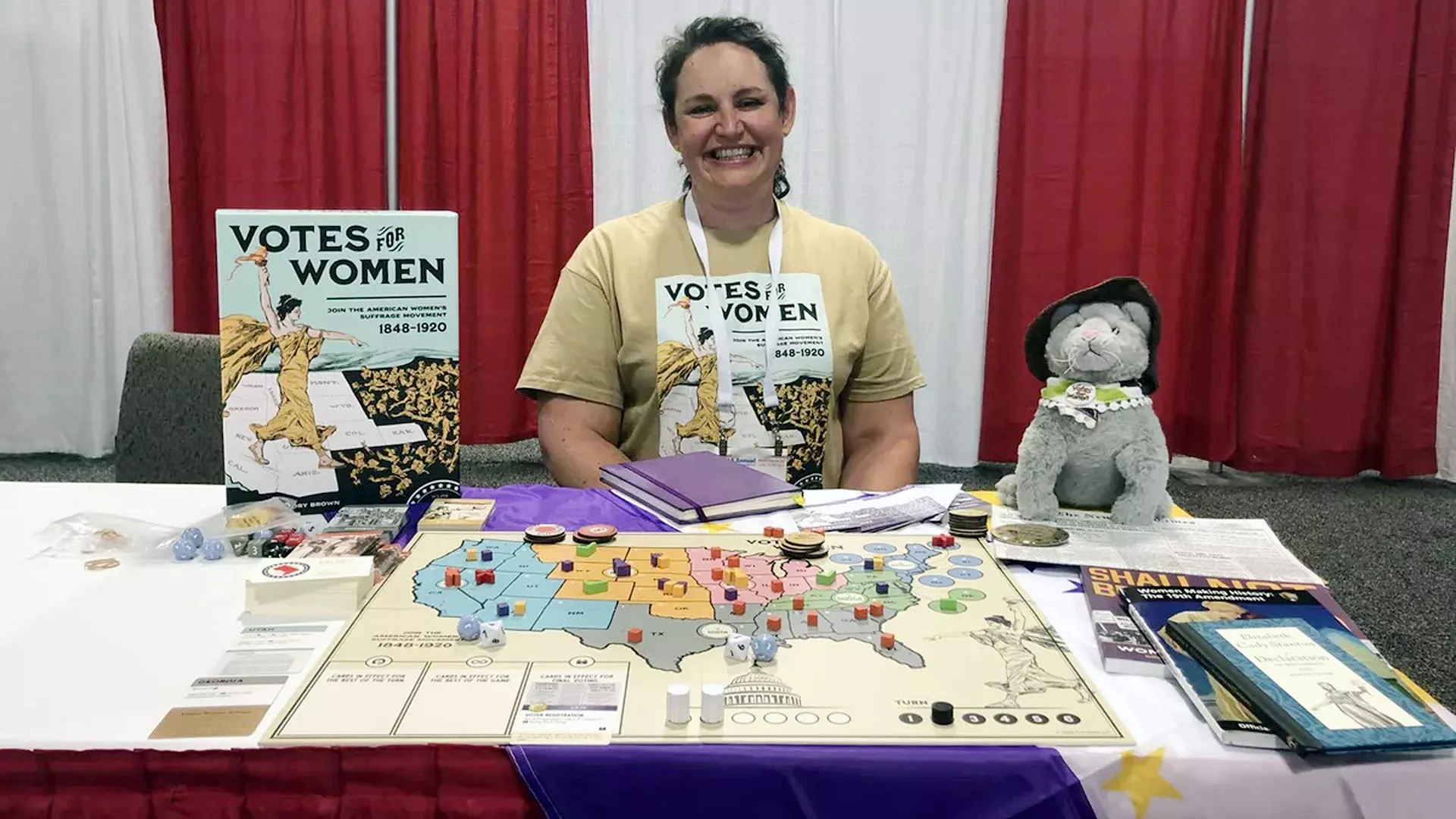Tory Brown sits at a table with a setup of the board game Votes for Women and a stuffed gray cat