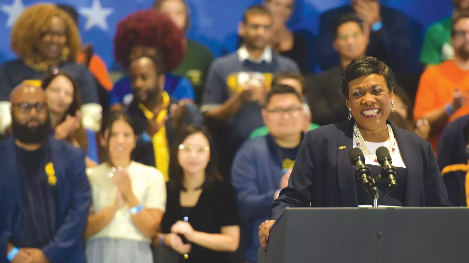 NEA President Becky Pringle smiling at the podium with a diverse group of NEA members behind her.