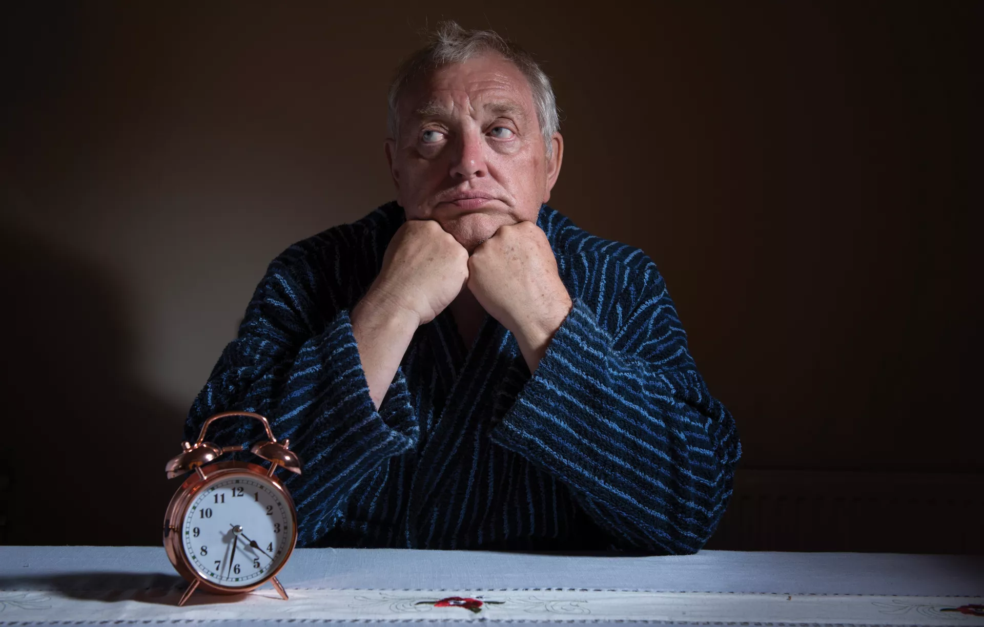 Older man in pajamas sitting at a table with an alarm clock on it, looking like he can't sleep.