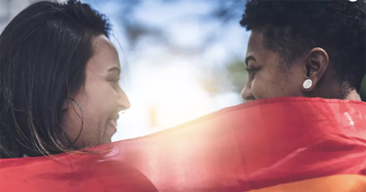 Two women facing each other smiling draped by a rainbow flag