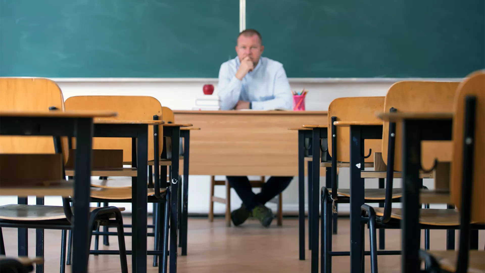 Stressed male teacher sitting at teacher's desk in otherwise empty classroom
