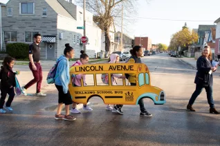 students and adults cross the street with a cut out of a bus that says "walking bus"