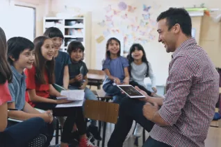 Man with iPad sits on a desk in front of a group of students.