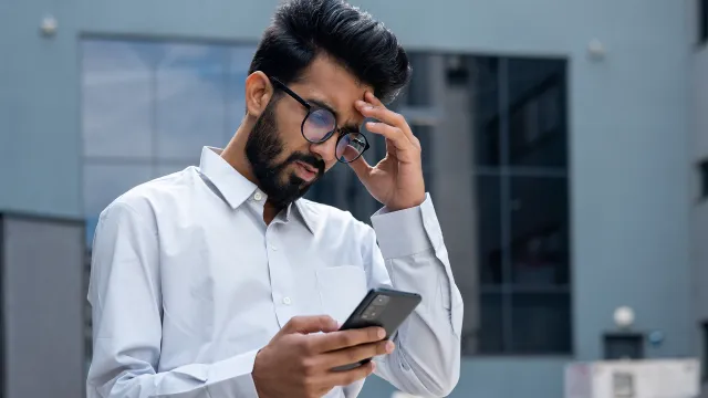 Latino man in white shirt and glasses looks at his phone with a worried expression