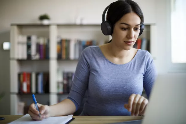 photo of a Latina woman wearing a blue shirt and headphones taking notes as she watches a webinar on her laptop