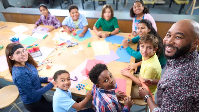 Students and teacher sit around a table and look up into the camera