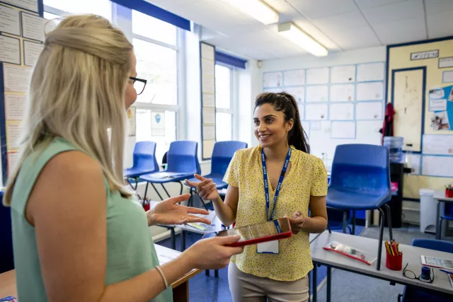 photo of blonde-haired educator speaking to a brown-haired educator in a classroom